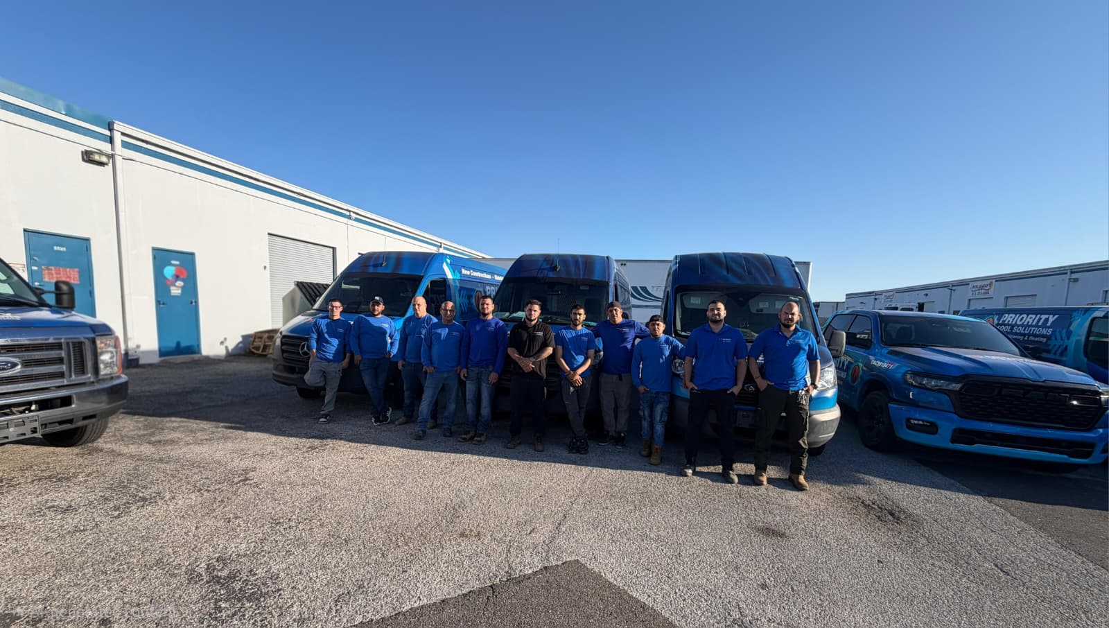Team of workers in blue uniforms stand in front of marked company vans and trucks on a sunny day.