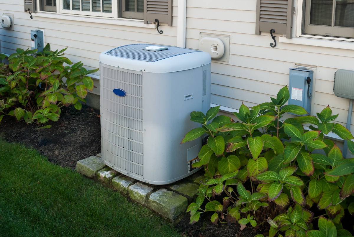 Outdoor air conditioning unit next to house with green plants.