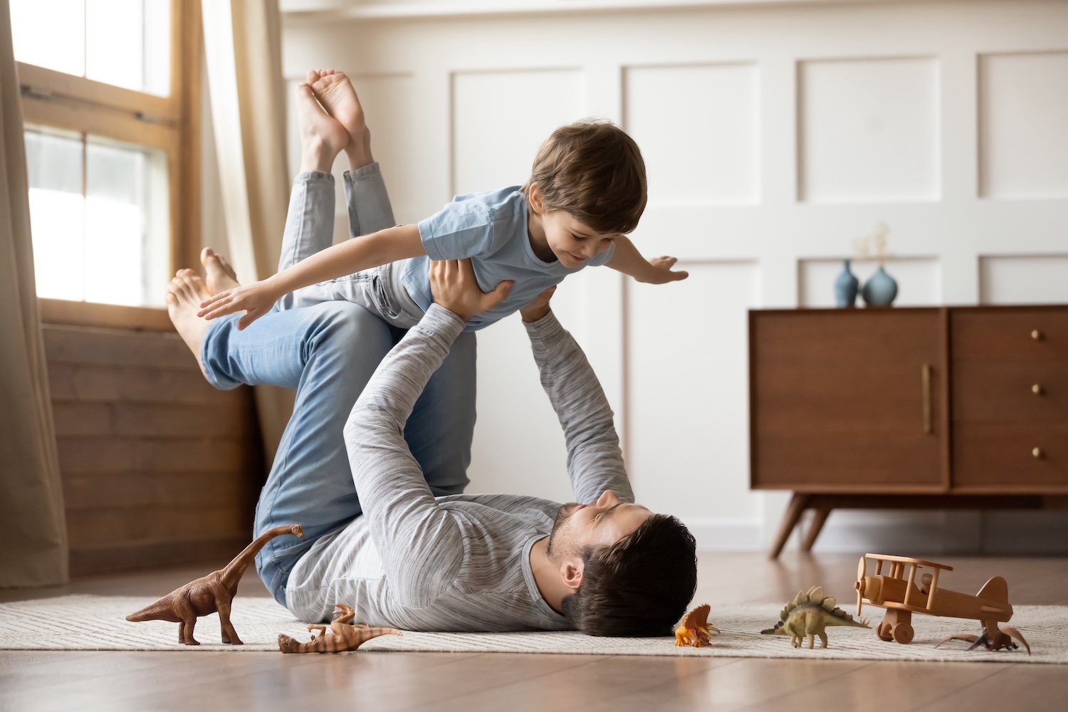 A person playfully lifting a child in a living room with toy dinosaurs and a plane.