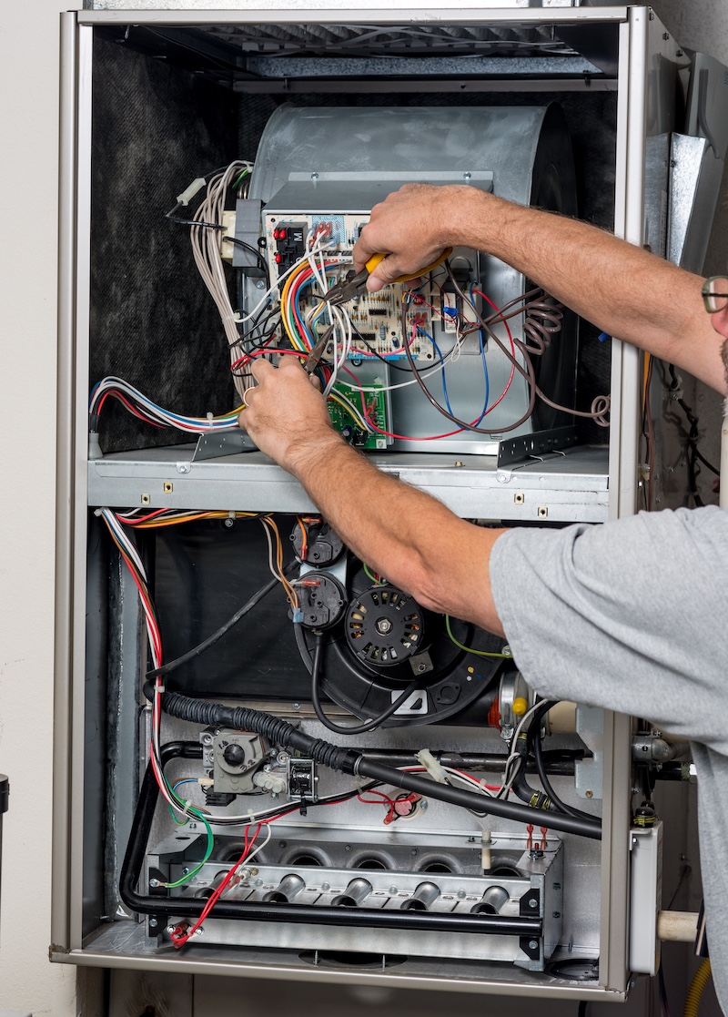 Technician repairing an HVAC furnace with tools and wires exposed.