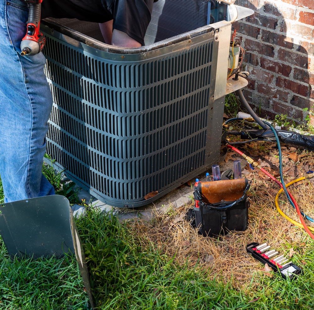 Technician repairing outdoor air conditioning unit with tools in grass.