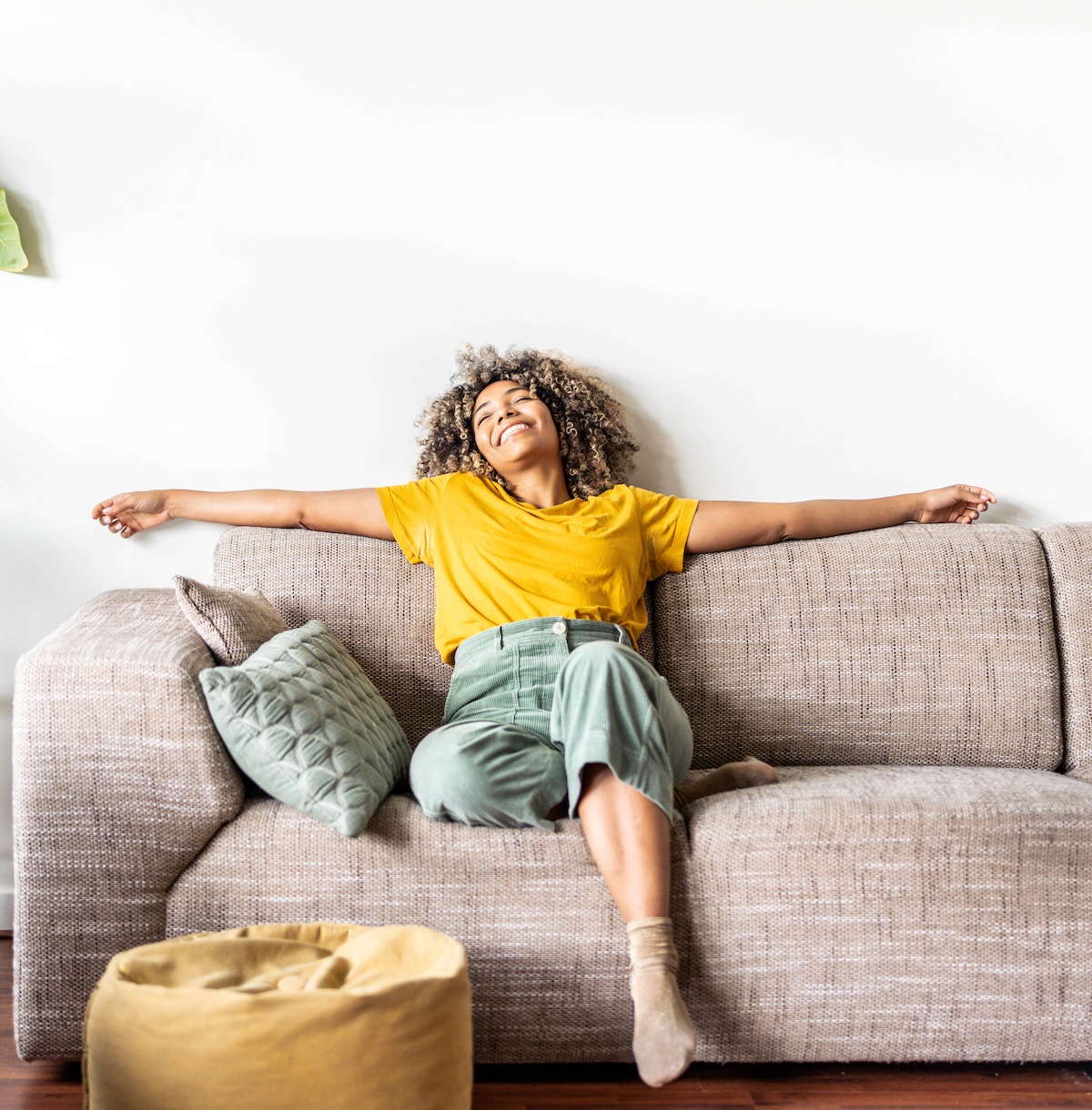 Woman in a yellow shirt relaxing on a couch with arms outstretched, smiling joyfully in a cozy living room.