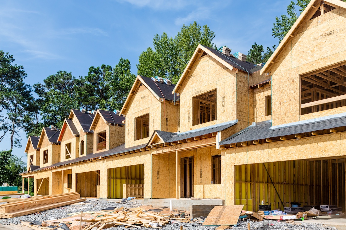 Row of wooden houses under construction with frames visible, surrounded by trees on a sunny day.
