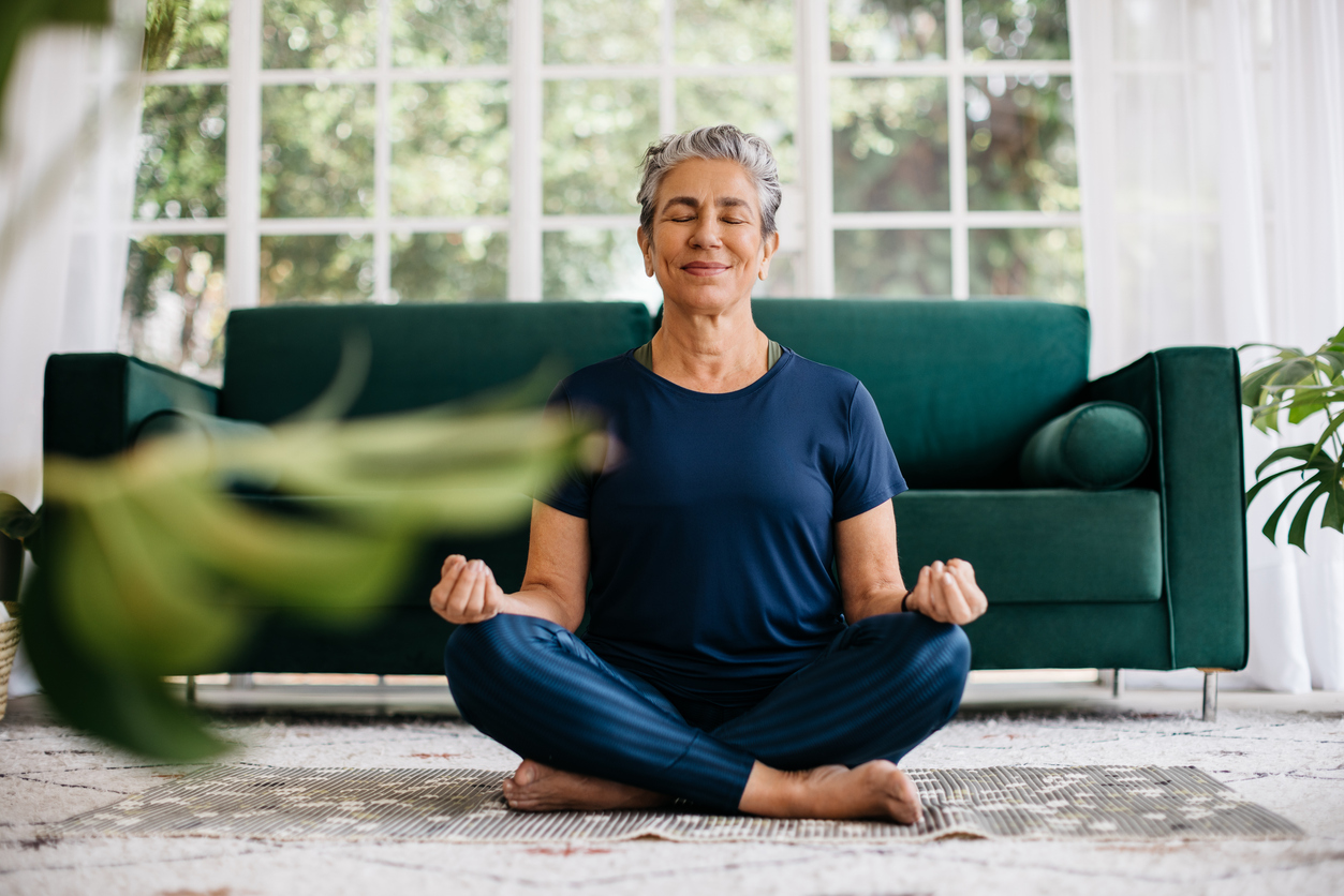 A person meditating in a bright living room, sitting cross-legged on a mat with a green couch in the background.