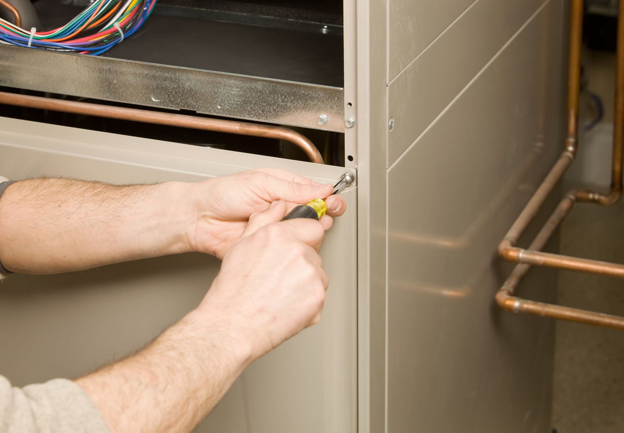 Person repairing a furnace with a screwdriver, focusing on metal casing and wiring.