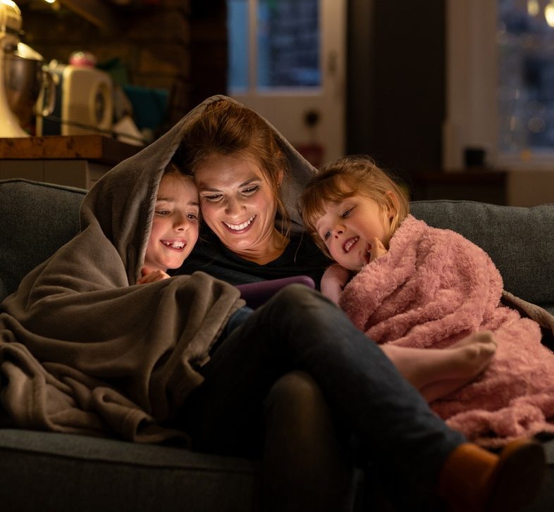 A woman and two children under blankets, smiling, while looking at a tablet on a cozy couch.