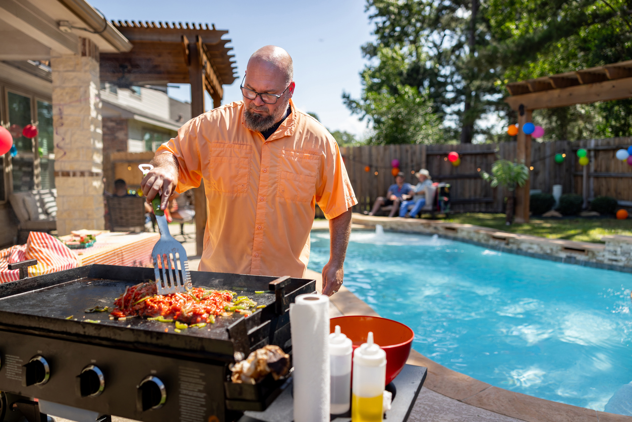 Man grilling vegetables by a poolside during a backyard summer party.