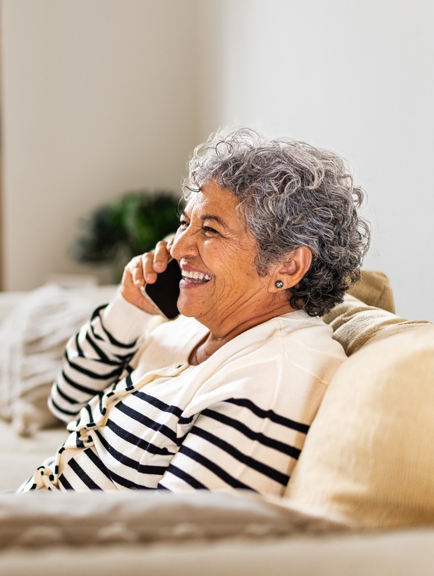 A smiling woman with curly gray hair on the phone, wearing a striped sweater, sitting on a couch.