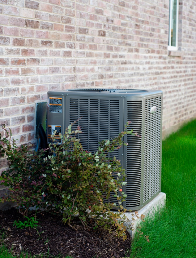 Outdoor air conditioning unit next to a brick wall with green plants.
