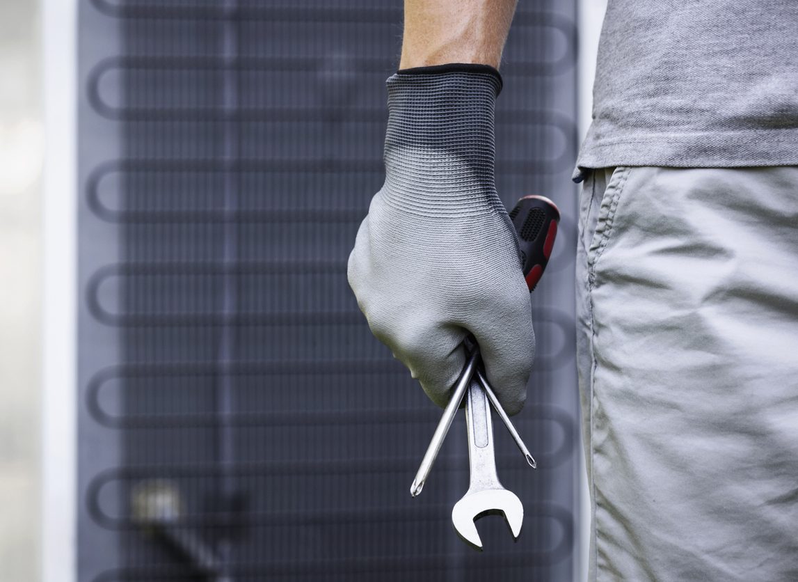 A gloved hand holding tools, standing near a large industrial unit.